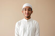 A young Muslim man, smiling and dressed in a white thobe and kufi, isolated on a beige background