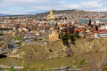 Wall Mural - Scenic cityscape of historical area of Tbilisi overlooking Holy Trinity Cathedral with golden dome in background and small Metekhi Church on steep bank of Mtkvari River on sunny spring day, Georgia