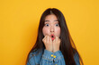 shocked chubby asian girl with little long hair in denim shirt with hand over lips over yellow background