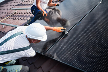 Wall Mural - Workers building photovoltaic solar panel system on rooftop of house. Close up of men technicians in helmets and gloves installing solar module with help of hex key outdoors. Renewable energy.