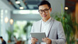 © DesiArt - Asian man looking into his tablet standing in office - ai generative