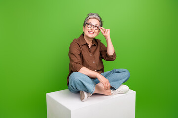 Poster - Full length photo of dreamy clever cheerful woman sit on cube touch eyewear look at offer empty space isolated on green color background