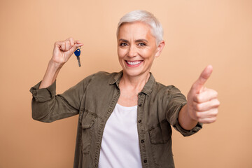 Poster - Photo portrait of lovely pensioner lady show thumb up hold key dressed stylish khaki garment isolated on beige color background