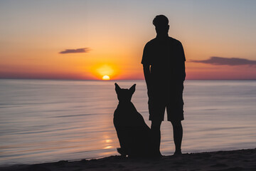 Silhouette of a man with a dog on the beach watching sunset close together. Owner and pet friendship concept