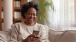 © BajimBa - A smiling, happy middle-aged black business woman in the office is sitting on the couch and looking at the phone she is holding in her hands