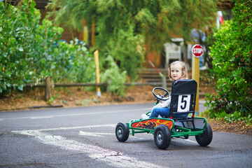 Preschooler girl driving pedal race car in amusement park with road surface marking