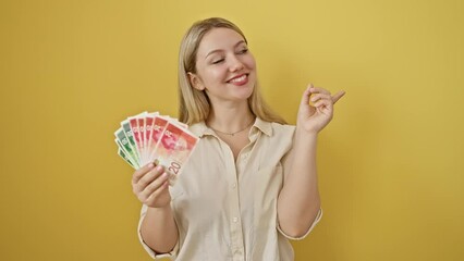 Sticker - Cheerful young blonde woman gleefully clutching israeli shekel banknotes, confidently pointing to the side with an optimistic smile on her face, set against a vivid yellow, isolated background