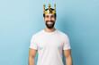 Portrait of man wearing white T-shirt and crown on head, smiling, concept of self confidence in success, self-motivation and dreams to be best. Indoor studio shot isolated on blue background.