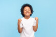 © khosrork - Portrait of extremely happy optimistic little boy with curly hair closed eyes clenched fists celebrating his success in school. Indoor studio shot isolated on blue background.