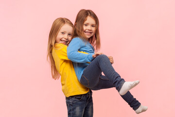 Portrait of two playful cute positive little girls having fun together, playing active games, expressing joy and happiness. Indoor studio shot isolated on pink background.