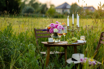 Romantic sunset wedding table decor for a couple with vintage chandelier, burning candles, snacks, white wine, fruits, ranunculi flowers. Summer vibes, field or meadow, outdoors event