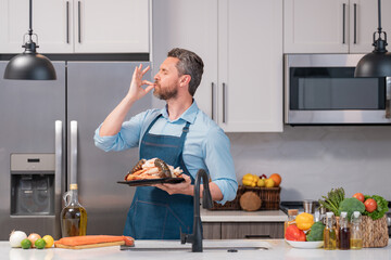 Poster - Man in cook apron preparing seafood at home modern kitchen. Man chefs with sign of perfect food. Chef man cooking, showing sign for delicious. Tasty delicious gesture by kissing fingers.