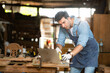 © Wosunan - Carpenter working on laptop in his workshop at a woodworking factory