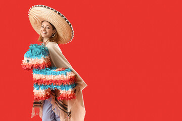 Wall Mural - Pretty young woman in sombrero and poncho with pinata on red background