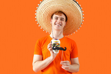 Canvas Print - Portrait of young Mexican man in sombrero, with painted skull and paper mustache on orange background. Celebration of Day of the Dead