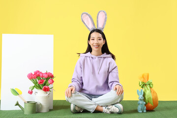 Sticker - Young Asian woman in bunny ears with Easter decor and blank poster sitting on grass against yellow background