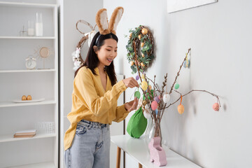 Sticker - Young Asian woman in bunny ears decorating willow branches with Easter eggs at home