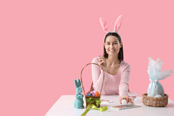 Canvas Print - Young woman in bunny ears with basket of Easter eggs and paints at table on pink background