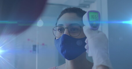 Poster - Image of glowing light trails over woman wearing face mask having temperature checked