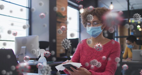 Poster - Image of covid 19 cells over woman in office disinfecting smartphone, wearing face mask