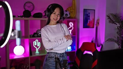 Poster - A cheerful young asian woman with headphones poses in a vibrant gaming room at night, exemplifying modern home entertainment.