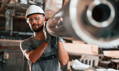 Moving big metal pipe. Young factory worker in grey uniform