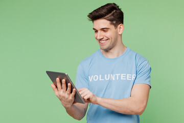 Sticker - Young smiling IT man wears blue t-shirt white title volunteer hold use digital tablet pc computer isolated on plain pastel green background. Voluntary free work assistance help charity grace concept.