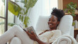 © Margo_Alexa - long shot of a 30 year old black woman, relaxing in a white reclining chair in her large bright living room, smiling while reading a magazine