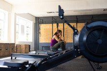 Side View Of Exhausted Biracial Young Woman Sitting On Rowing Machine In Health Club, Copy Space