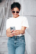 © halayalex - Young beautiful smiling hipster woman in trendy summer casual clothes. Carefree woman posing in the street. Positive model holds mobile phone, looks at cellphone screen, uses smartphone apps