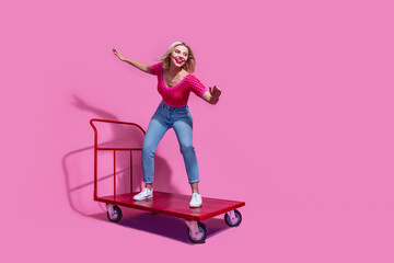 Wall Mural - Full length photo of eccentric lovely woman riding on shopping cart to empty space look at promo empty space isolated on pink background