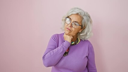 Poster - Pensive mature woman in glasses, grey-haired, standing over pink isolated background. cheerfully smiling, hand to chin, deep in thought, doubting.