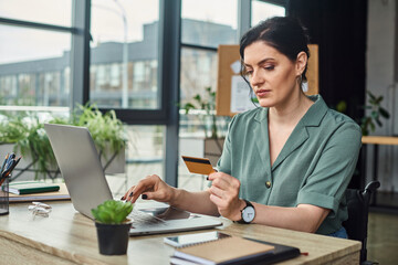 good looking dedicated woman in casual attire in wheelchair looking at credit card while at laptop