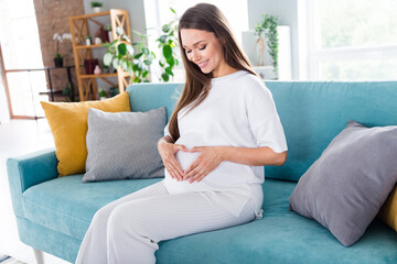 Wall Mural - Photo of lovely dreamy cute gentle mama pregnant girl dressed white pajama sitting on sofa touching belly waiting baby flat indoors