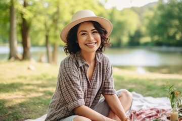 Wall Mural - Beautiful young woman in hat sitting on blanket in park and smiling