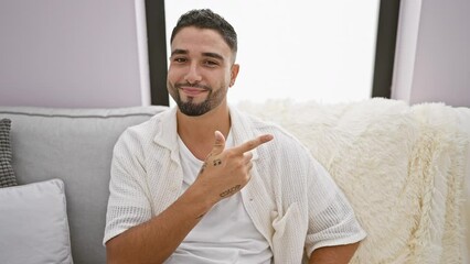 Wall Mural - Cheerful young arab man happily pointing up with a natural smile on his face, sitting on the sofa at home, presenting something on the side.