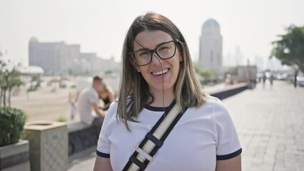 Wall Mural - Smiling woman wearing glasses in urban doha, featuring skyscrapers, sunny weather, and casual style.