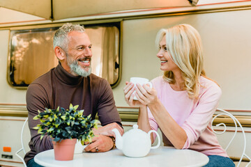 Wall Mural - Joyful senior european couple spouses husband and wife sitting at the table and drinking tea near the van. Traveling together caravanning by camper wheel motor home trailer