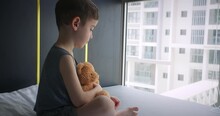 Little Boy Upset Child Of Preschool Age Sits Near Window On His Bed, Holds His Plush Bear In His Hands,looks Out Window Into Street.Violence Against Children In Family.Sad Kid.Child's Fear Loneliness.
