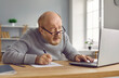 Senior man in glasses looking at laptop computer screen. Portrait of elderly man wearing gray knitted pullover browsing internet and making notes on paper while sitting at table at home