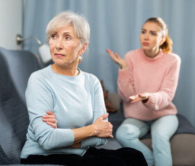 Wall Mural - Sad elderly woman having quarrel with her daughter. She's sitting on sofa and listening her daughter swearing.