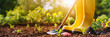 front view of gardening tools and a pair of yellow wellingtons outside on soil with plants, sunny weather