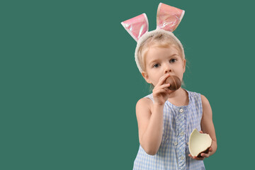 Wall Mural - Cute little girl in bunny ears eating chocolate egg on green background. Easter celebration