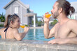Happy cheerful Caucasian white people couple lying on the beach bed beside swimming pool and drinking an orange juice together.