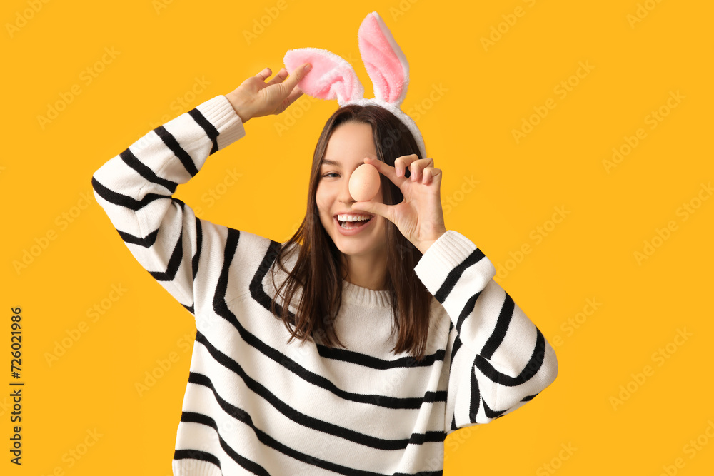 Pretty young woman with bunny ears and Easter egg on yellow background