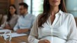 © mariiaplo - Close-up of a confident pregnant businesswoman in white shirt at a meeting, colleagues blurred