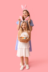 Wall Mural - Young woman and her daughter in bunny ears holding basket of Easter cake with painted eggs on pink background
