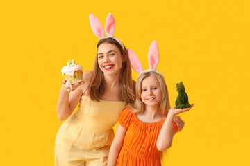 Poster - Cute little girl with rabbit and her mother in bunny ears holding Easter cake on yellow background