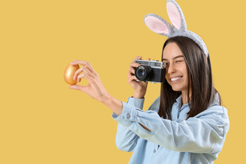 Wall Mural - Young woman in bunny ears with photo camera and Easter egg on yellow background