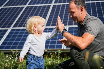 Wall Mural - Father and his little son giving each other high-five on background of solar panels. Young father glad spending time with his child. Happy family of two on background of solar station.